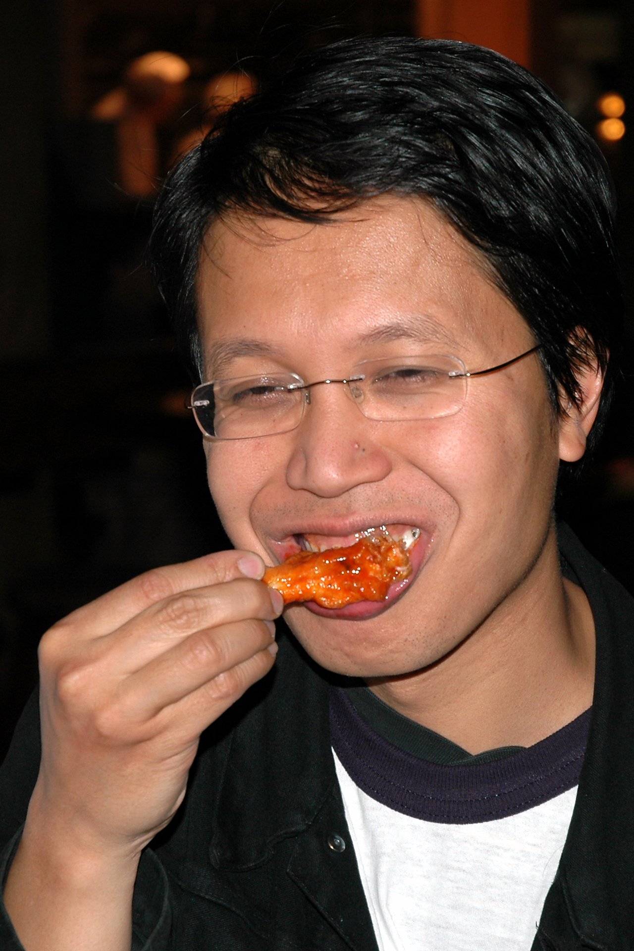 A person wearing glasses eats a saucy chicken wing, holding it close to their mouth while biting down.
