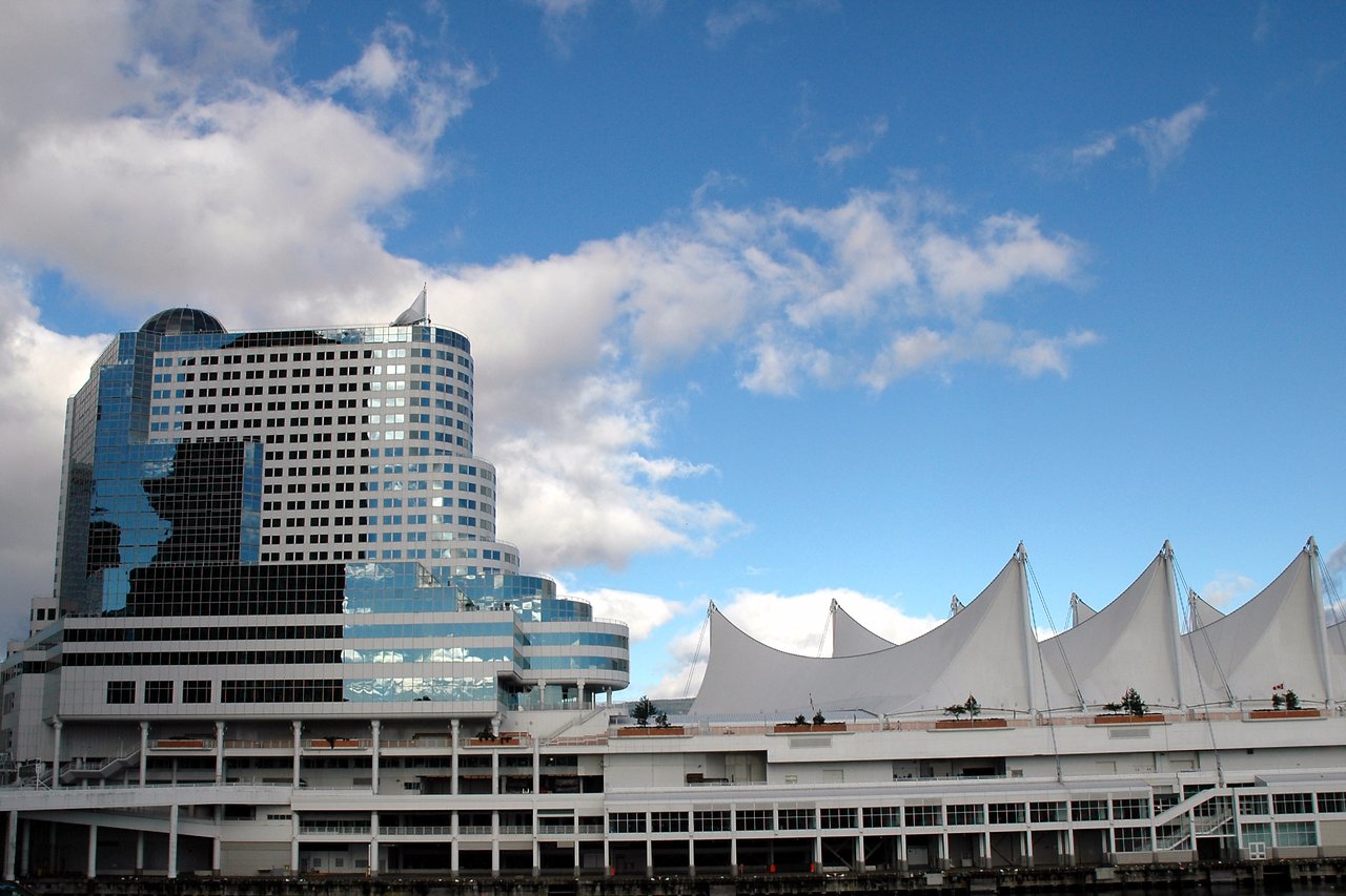 A waterfront conference venue with a modern glass building and white sail-like roof structures under a partly cloudy sky.