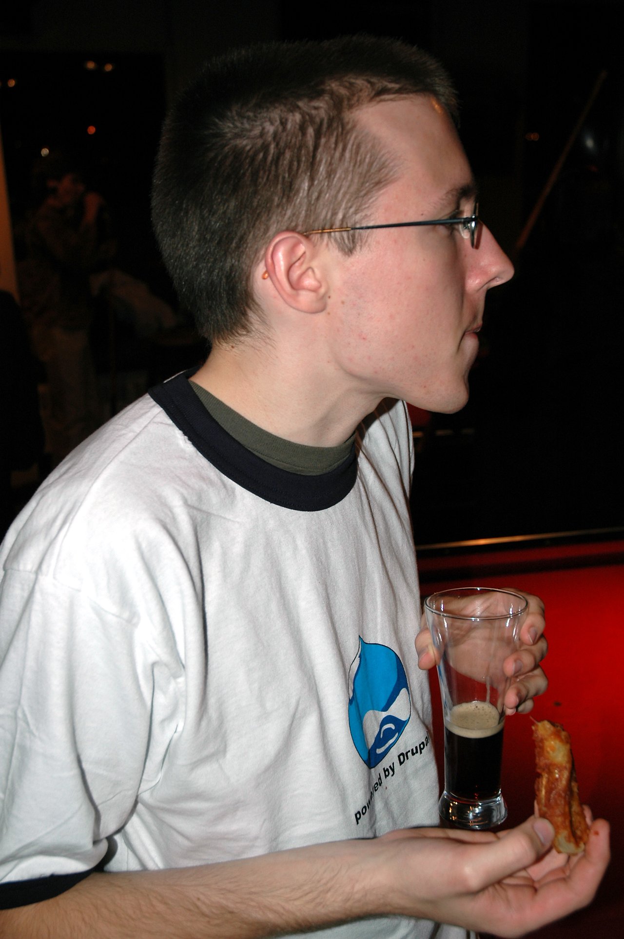 A person wearing a Drupal t-shirt holds a glass of dark beer and a piece of pizza in a pub.