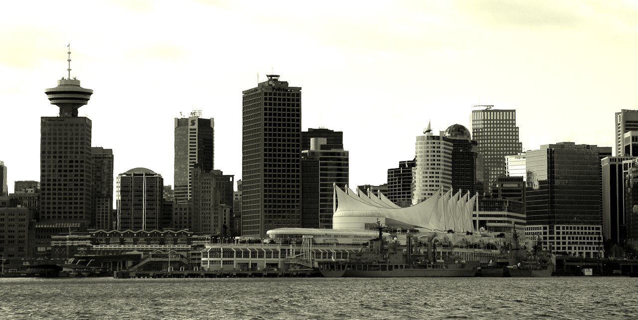 Vancouver skyline with high-rise buildings, Canada Place's white sails, and docked ships, viewed from across the water.