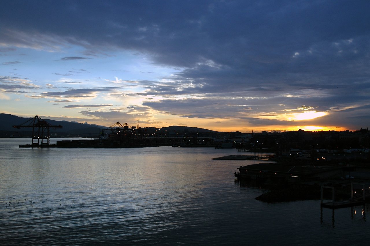 The sun sets over Vancouver's harbor, casting light on the water with silhouetted cranes and buildings in the background.