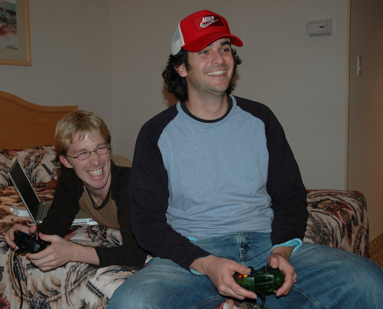 Two people playing Xbox in a hotel room, one laughing while the other smiles and focuses on the game.