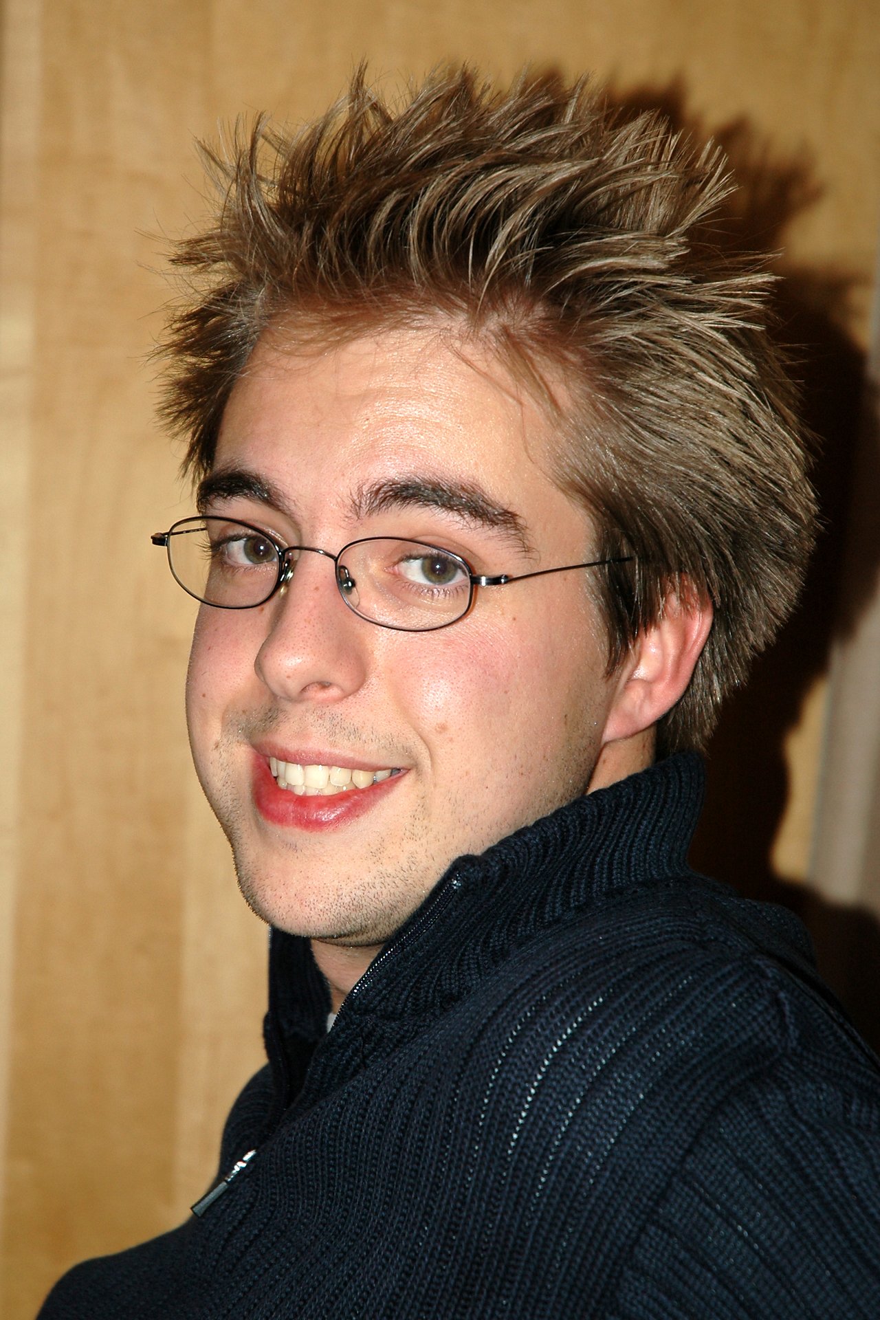 A person with glasses and spiky hair smiles at the camera, wearing a dark sweater indoors.
