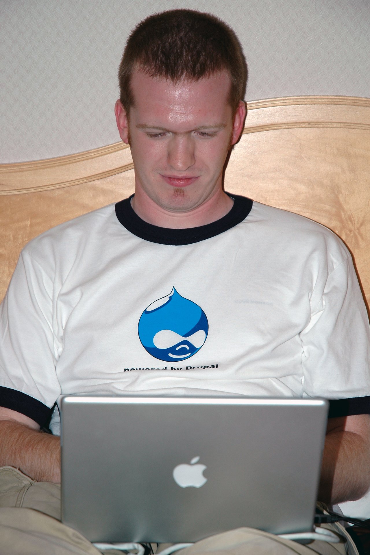 A man wearing a Drupal t-shirt sits on a bed, focused on working on his Apple laptop.