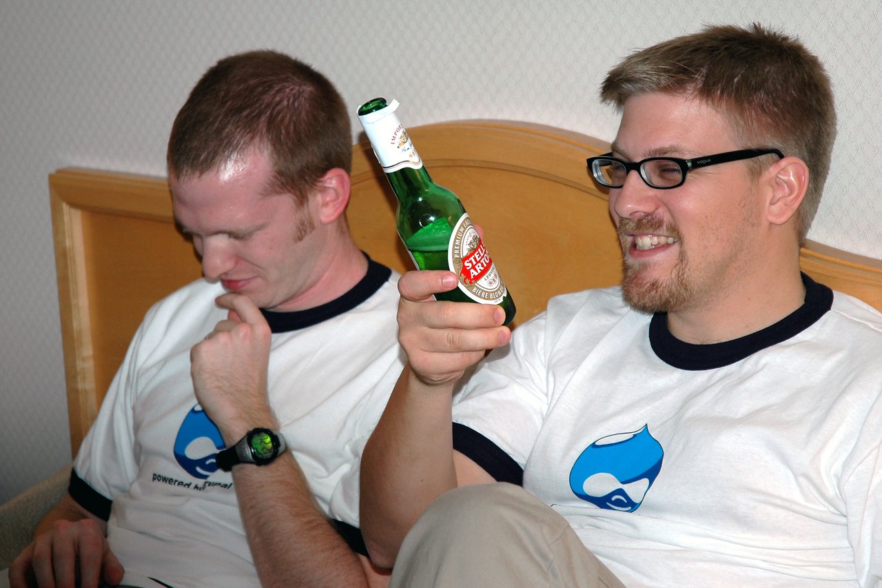 Two men in matching white shirts sit on a bed.