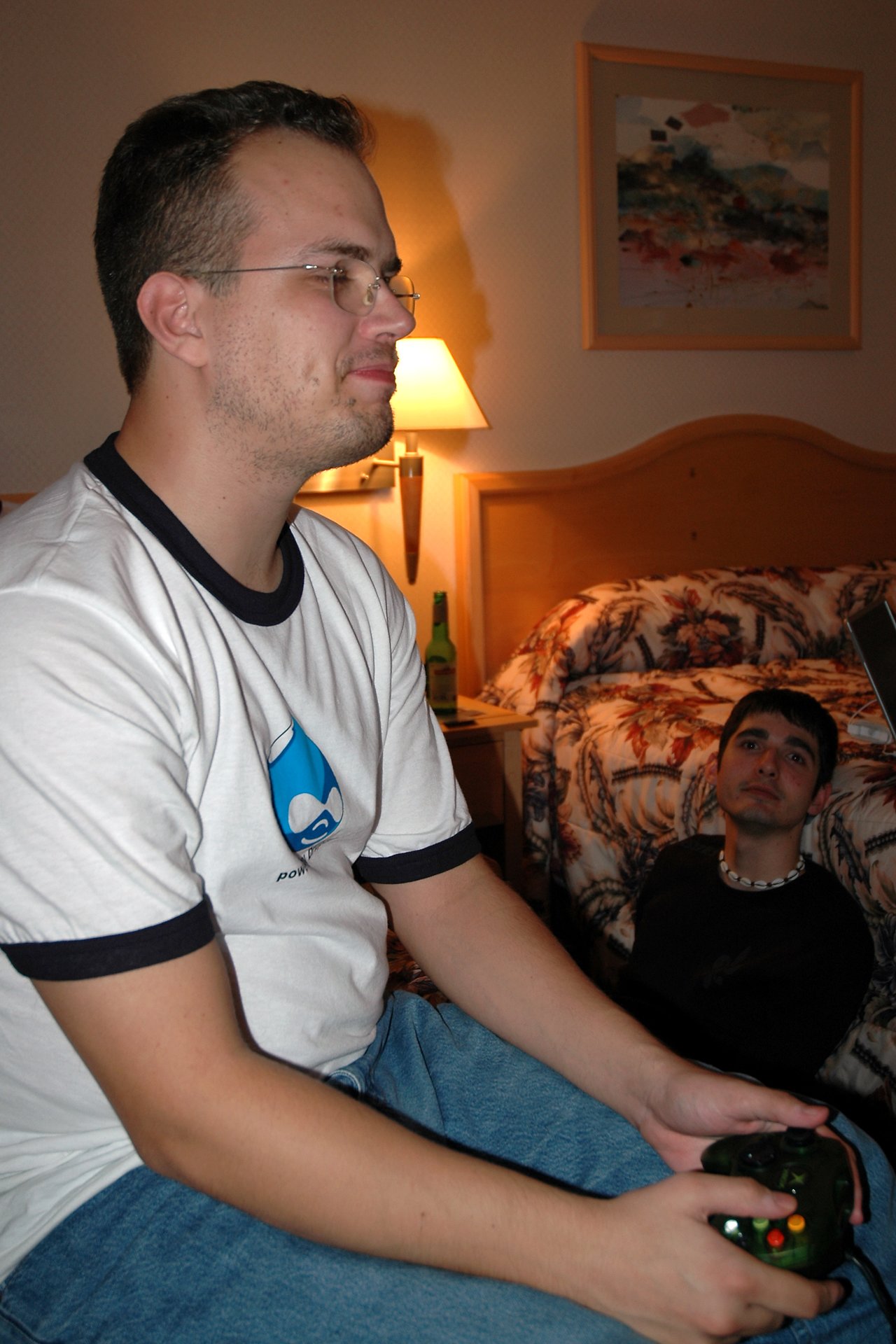 A man in a white shirt plays Xbox while another man sits on the floor watching.