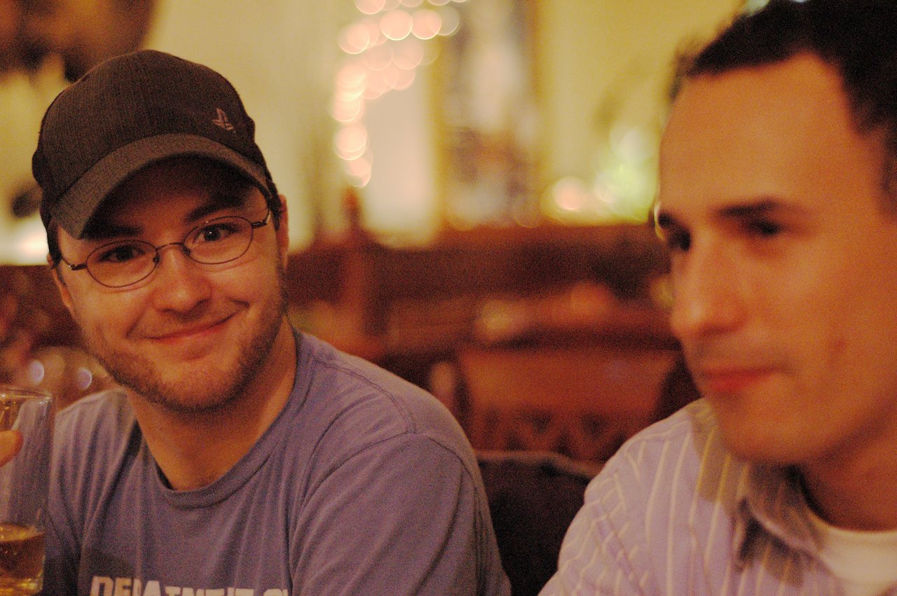 A man wearing a cap and glasses smiles while holding a drink, with another man blurred in the foreground.