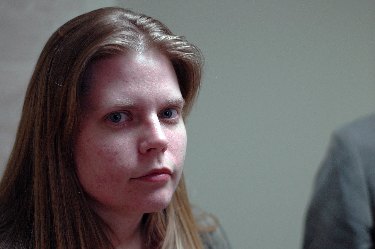 A person with long hair looks directly at the camera with a neutral expression in an indoor setting.