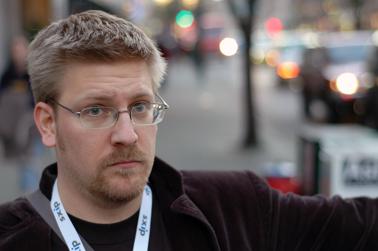 A man with glasses and a lanyard looks at the camera with a serious expression on a busy street.
