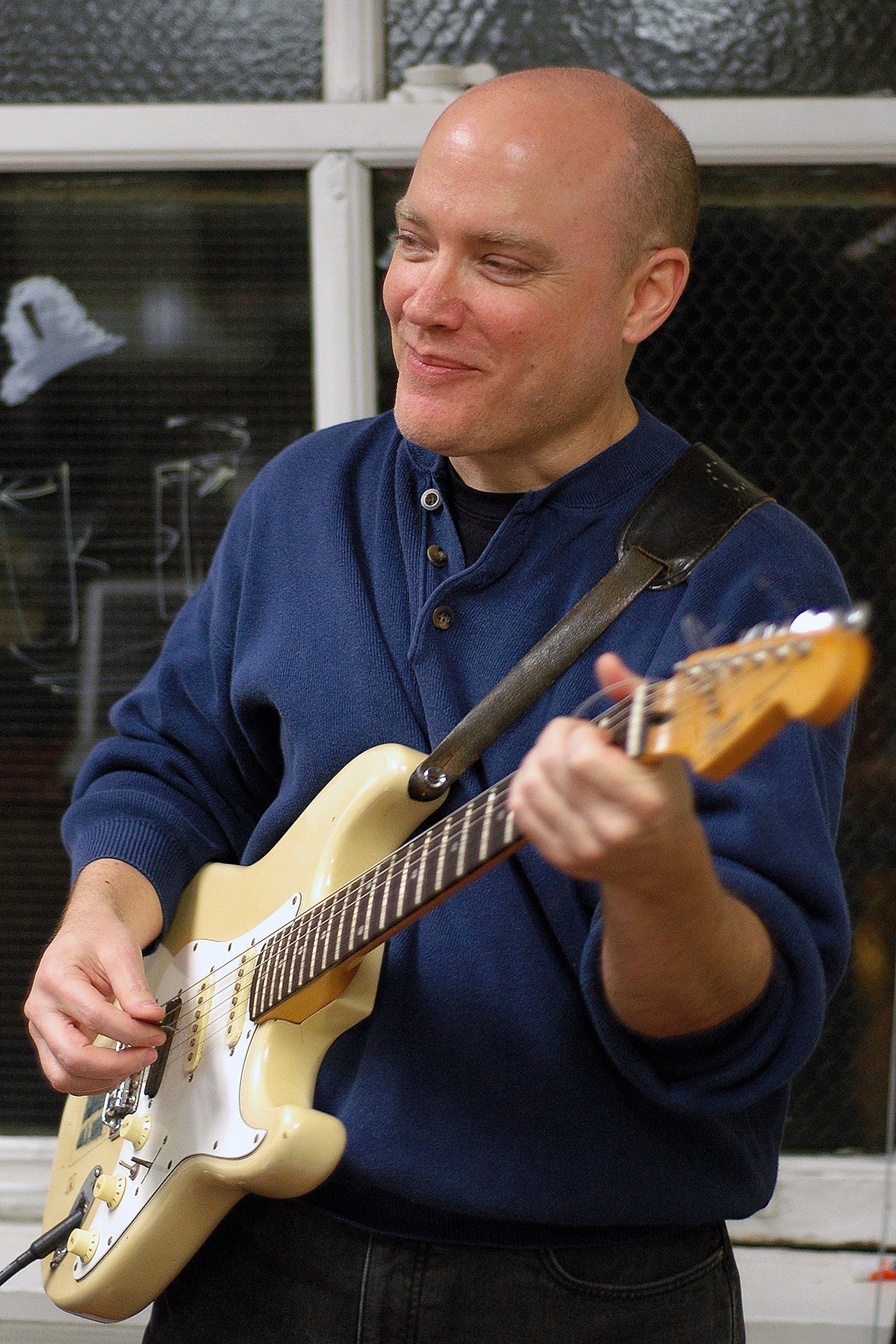 A man in a blue sweater plays an electric guitar and smiles.
