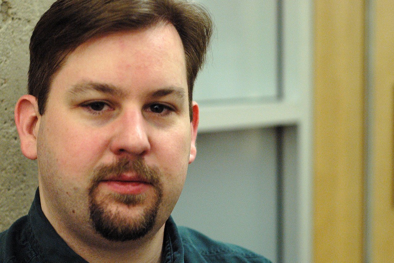 A man with short brown hair and a goatee looks directly at the camera in an indoor setting.