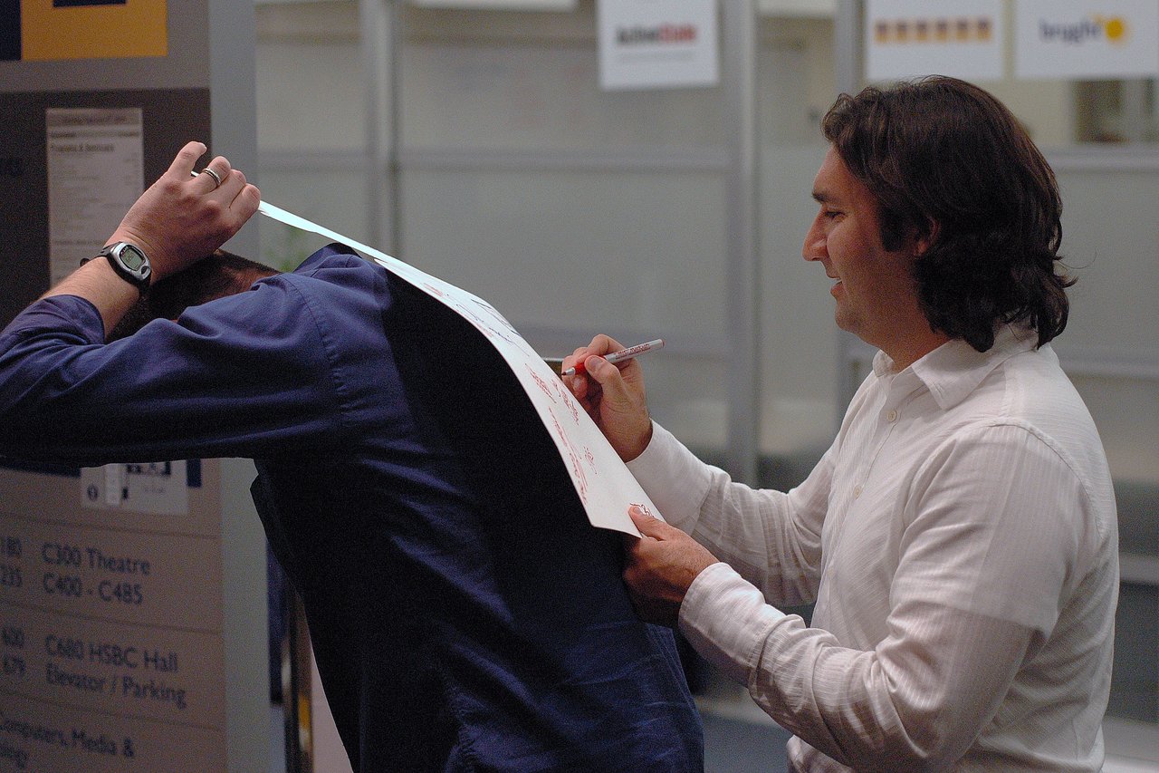 A man in a white shirt writes on a large sheet of paper resting on another person's bent back.