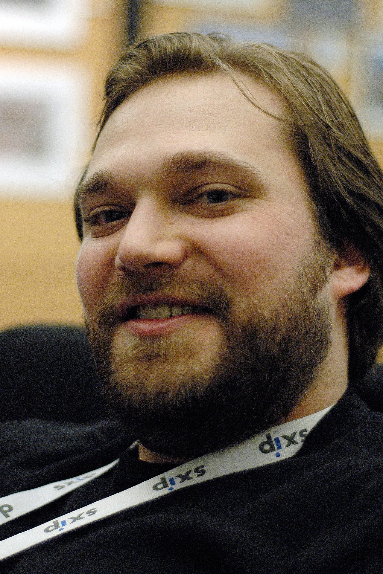 A bearded man wearing a lanyard smiles slightly while looking at the camera in an indoor setting.