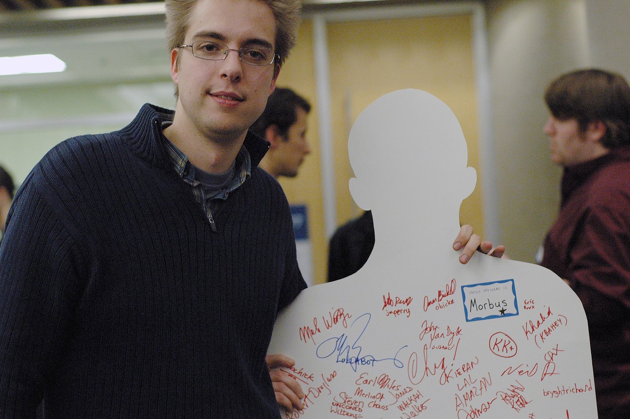 A person in a dark sweater holds a white cardboard cutout labeled "Morbus", covered in handwritten signatures.