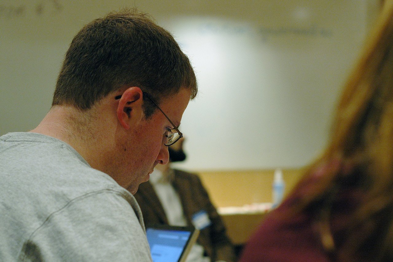 A man wearing glasses and a gray shirt is focused on something in front of him at a conference.
