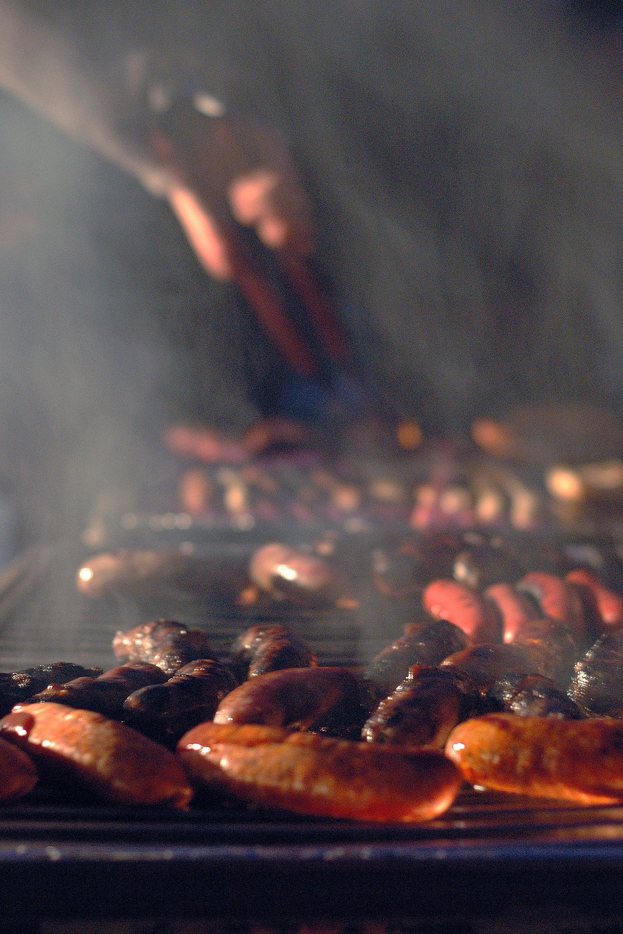 Sausages and other meats grilling at night, with a hand using tongs to turn them.
