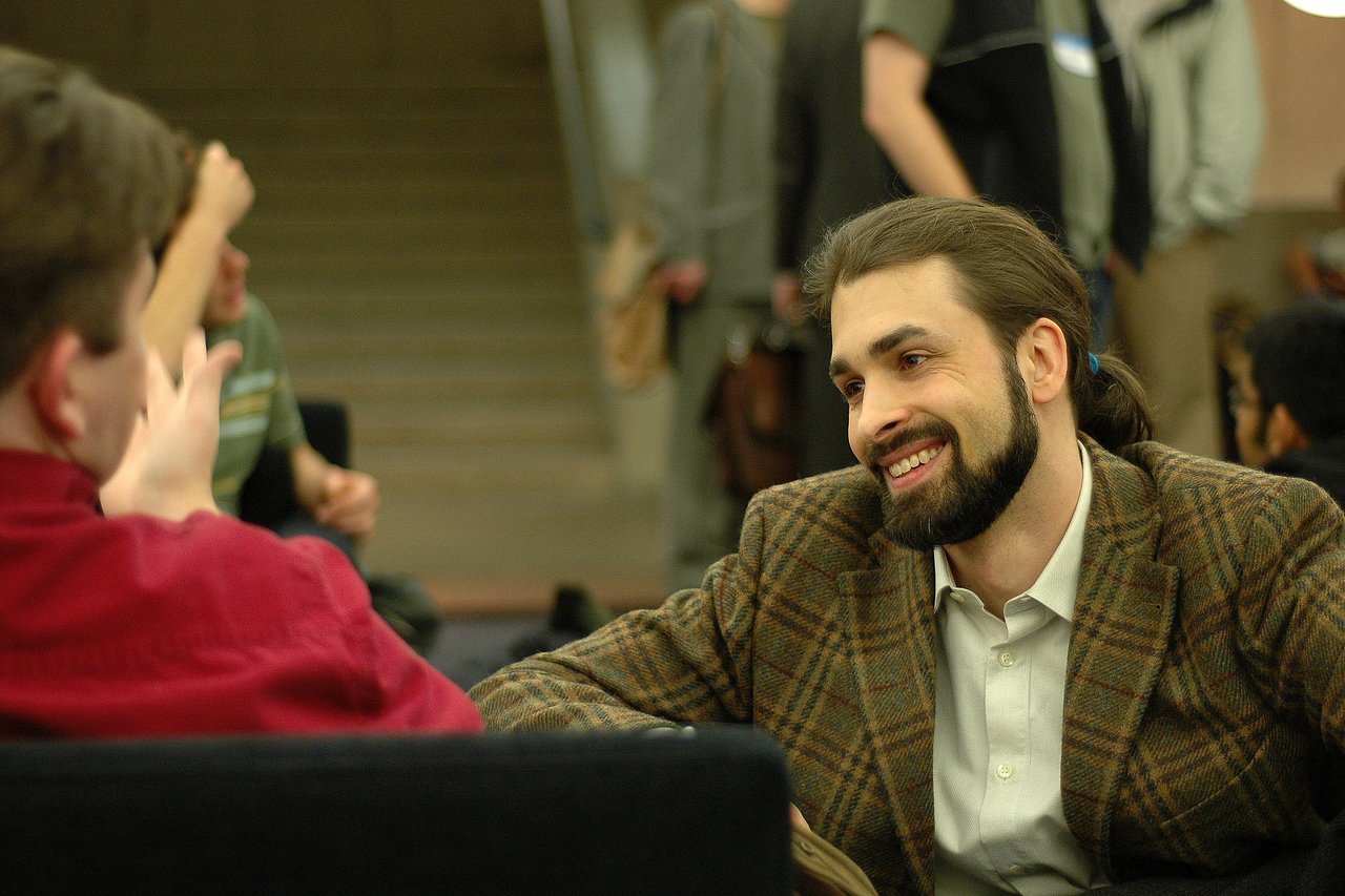 A man in a plaid jacket smiles while talking to another person wearing a red shirt.