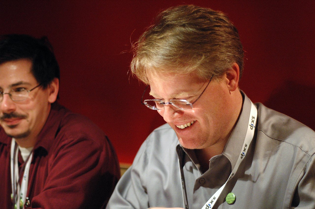 A man wearing glasses and a lanyard smiles while engaged in conversation at an event.