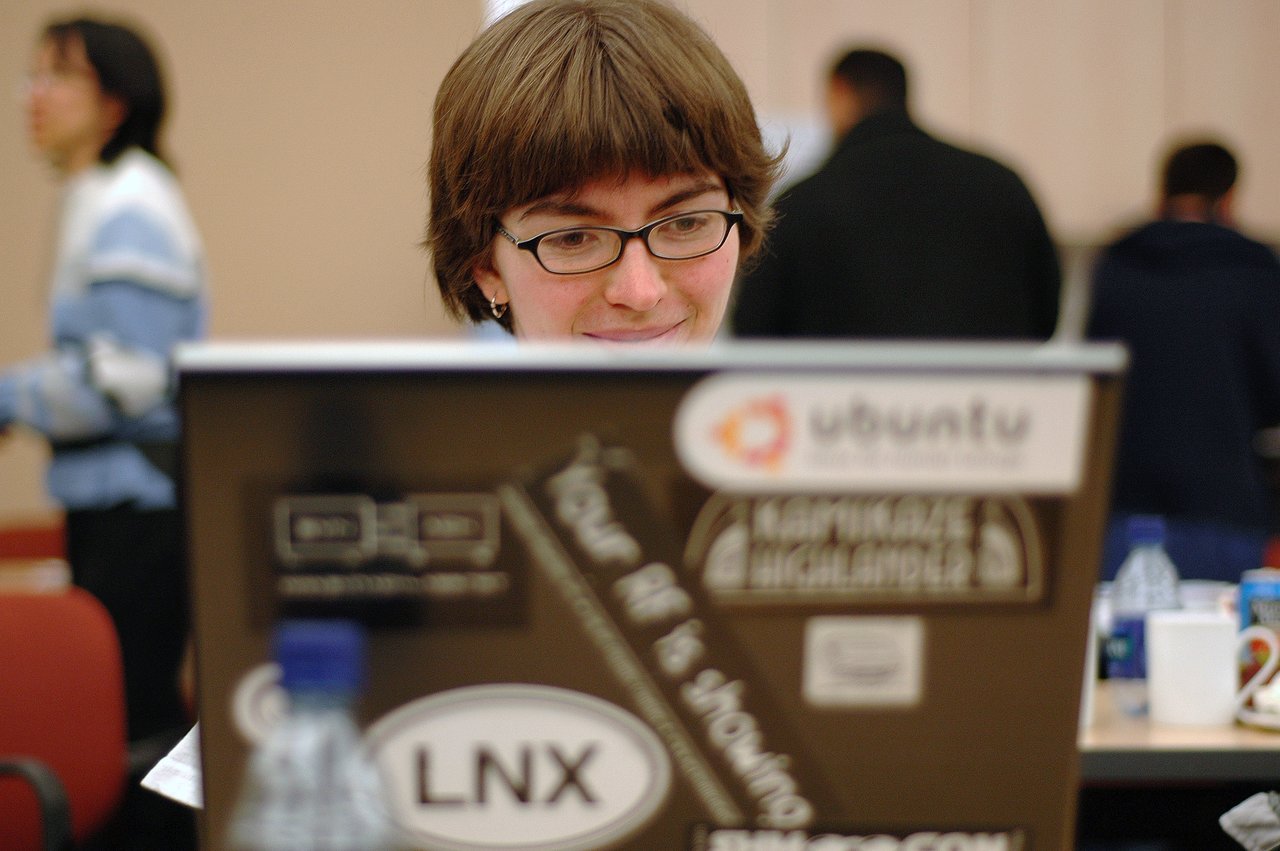 A person with glasses smiles while looking at a laptop covered in stickers, including an Ubuntu logo.