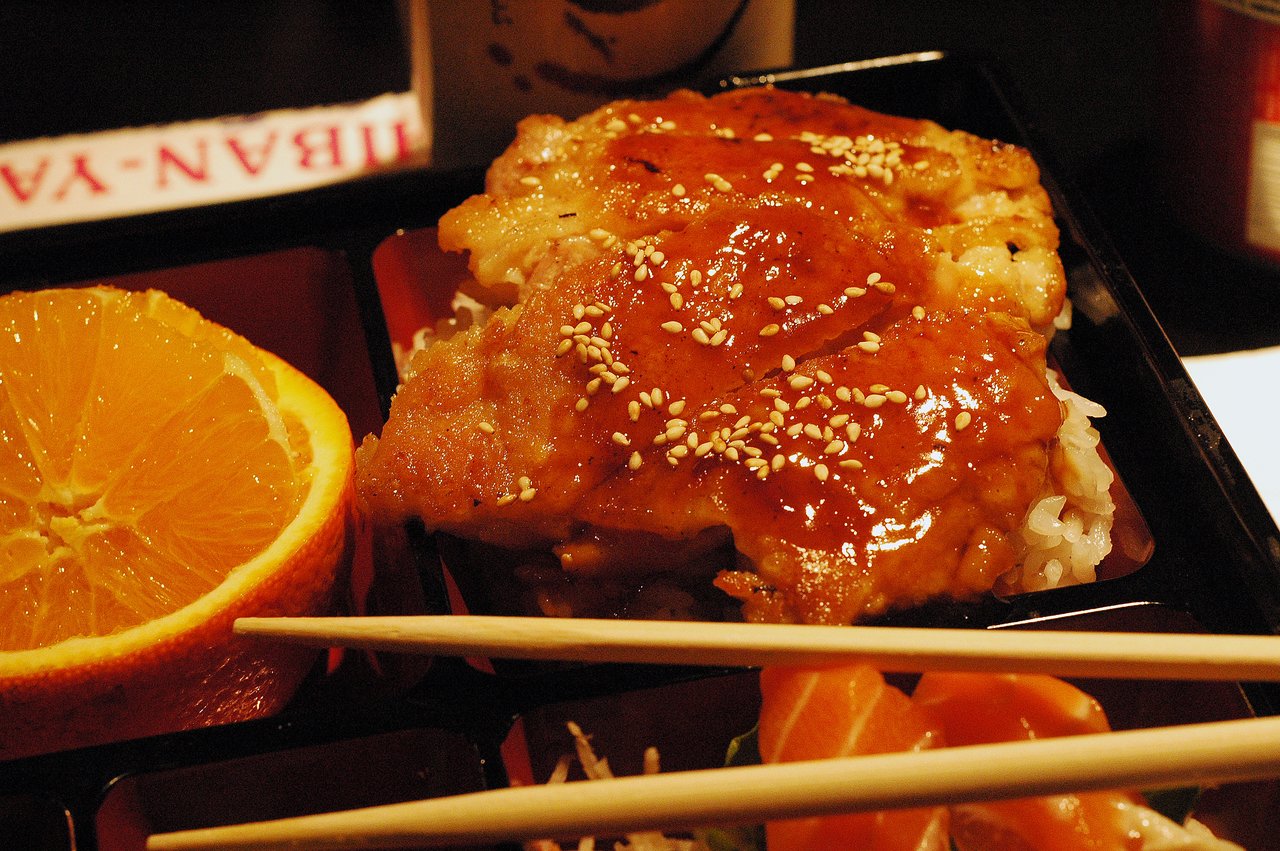 A sushi bento box with glazed fish, sesame seeds, orange slices, and chopsticks resting on top.
