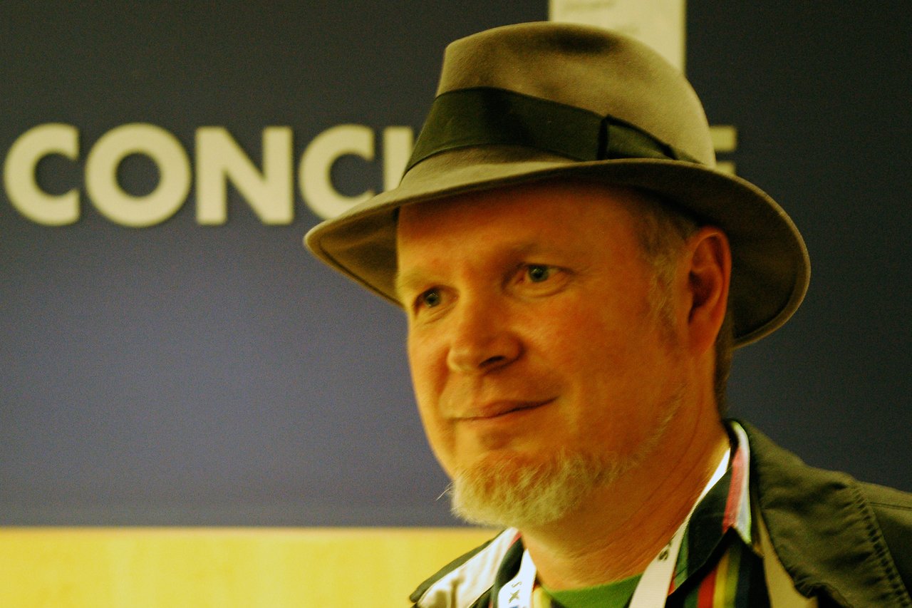 A man wearing a hat and a lanyard stands indoors, looking slightly to the side with a neutral expression.