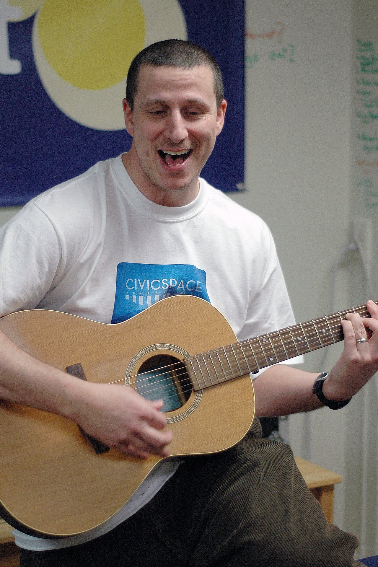 A man wearing a CivicSpace t-shirt plays an acoustic guitar and sings with an expressive face indoors.