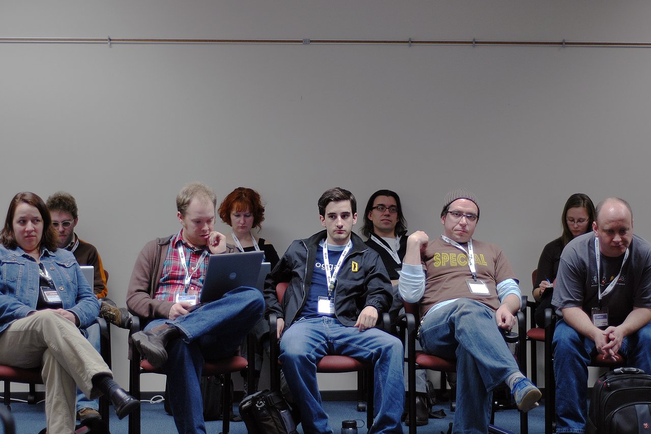 A group of people sitting in a room, some using laptops, with one person in the center looking forward.