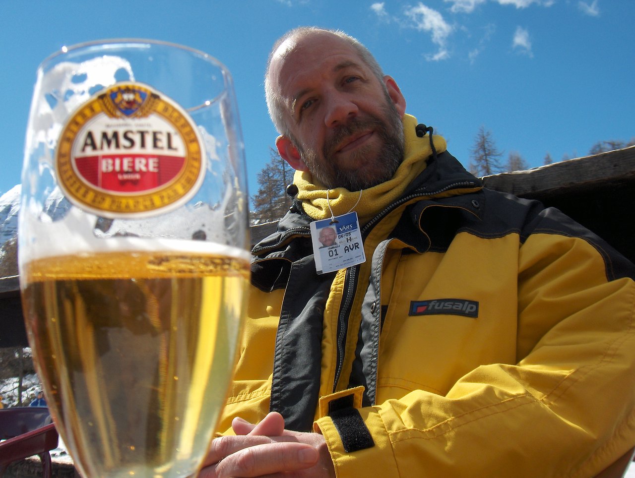 A man in a yellow jacket sits outdoors, looking at the camera, with a glass of beer in front.