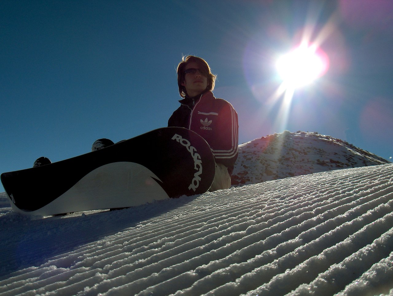 A person wearing sunglasses and a jacket sits on a snowy slope with a snowboard, facing the sun.