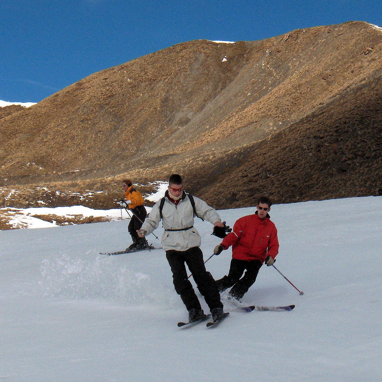 Two people skiing downhill, one in a white jacket and the other in red, with a third skier behind them.