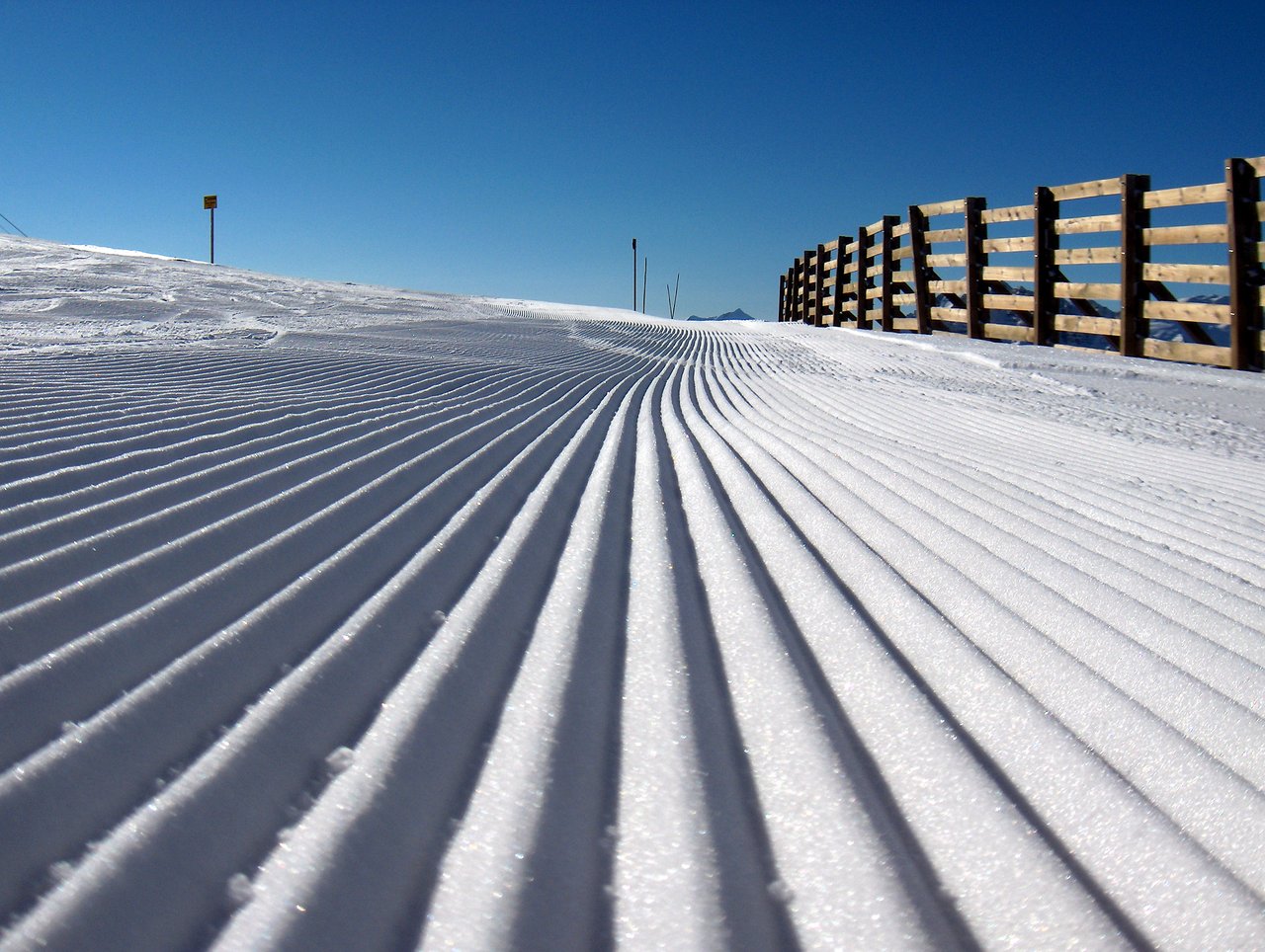 Freshly groomed snow with parallel ridges on a ski slope, bordered by a wooden fence under a clear blue sky.