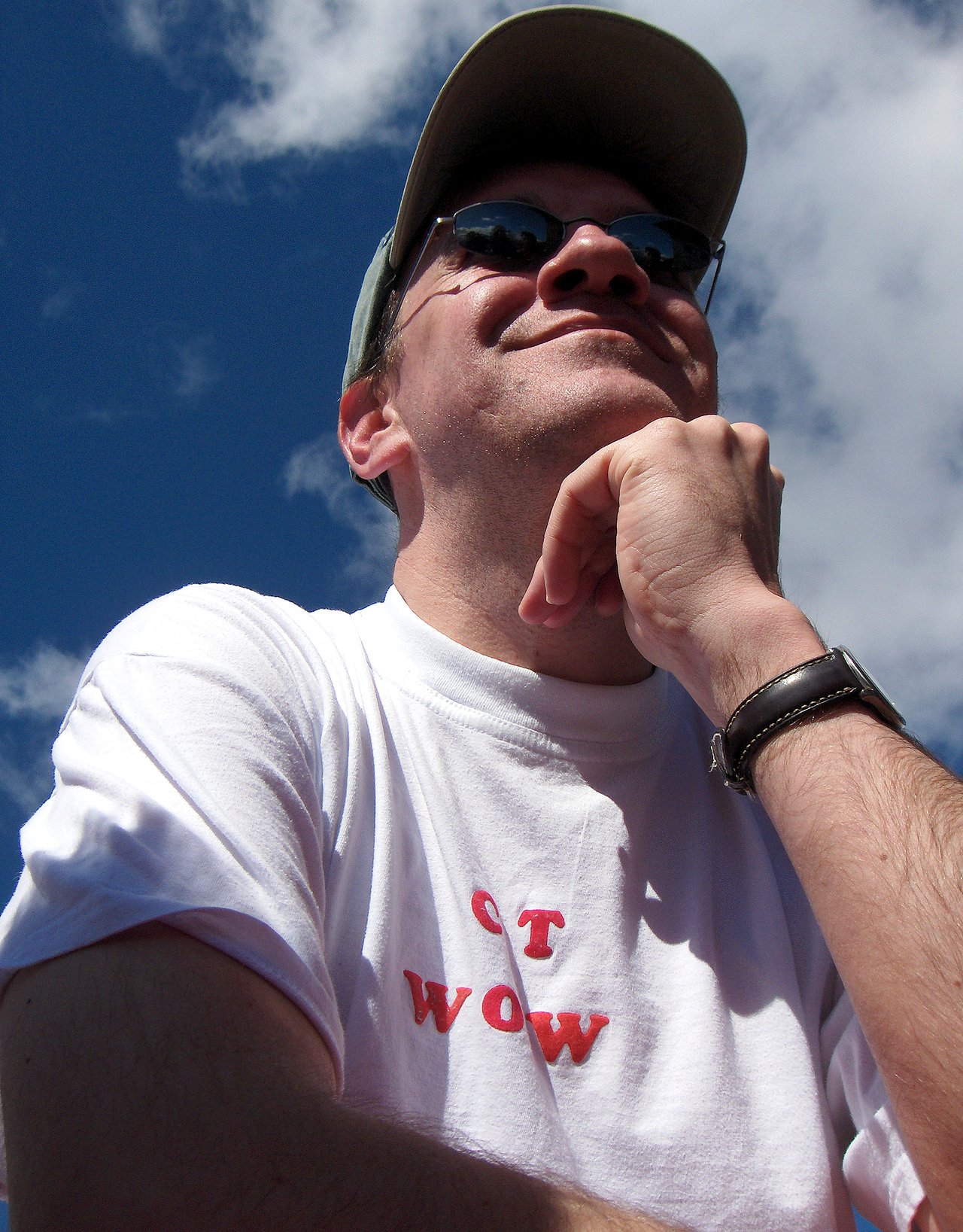 A man wearing sunglasses and a cap looks up thoughtfully, resting his chin on his hand.