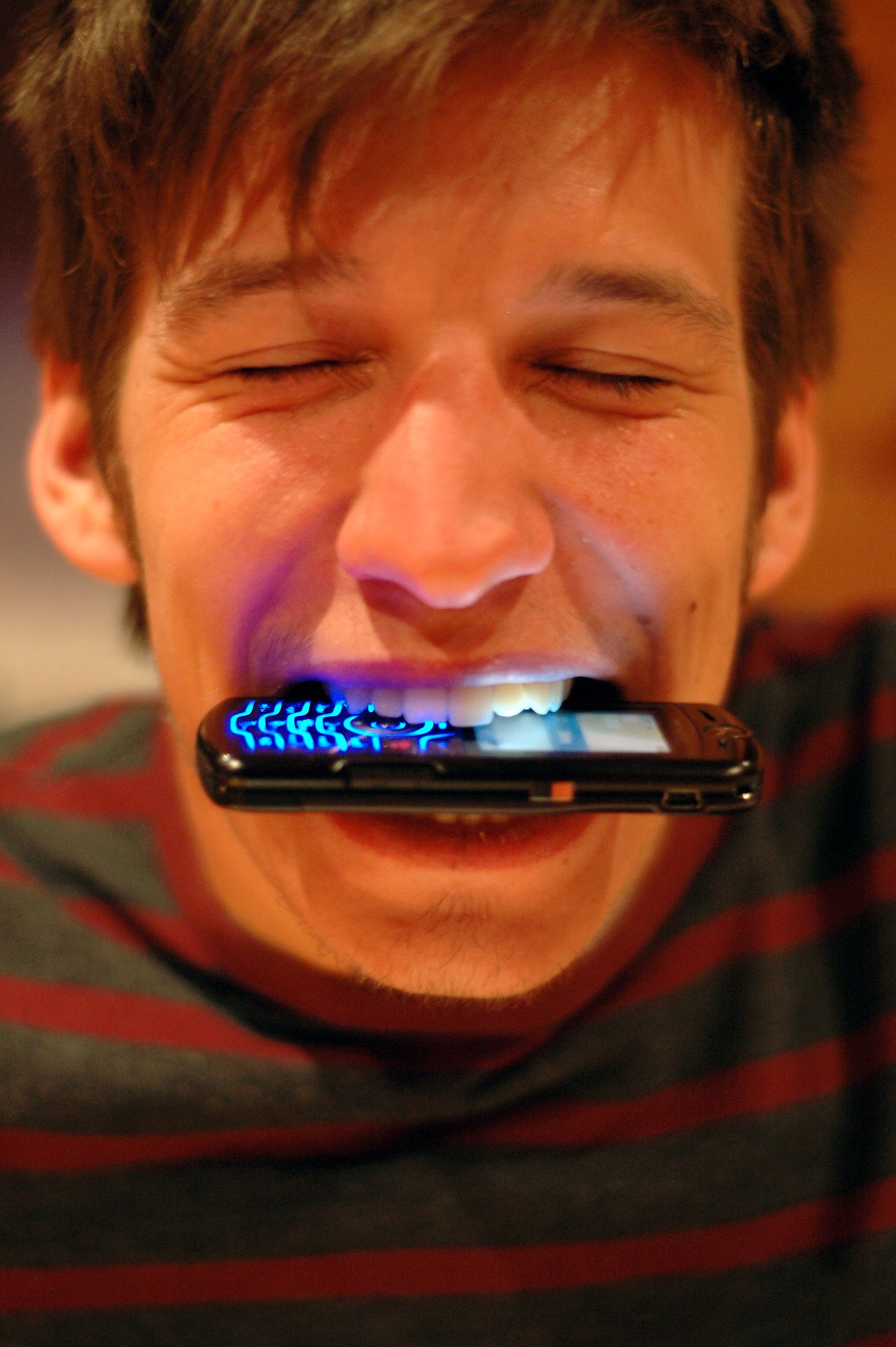 A person with closed eyes holds a glowing flip phone between their teeth, showing its illuminated keypad.
