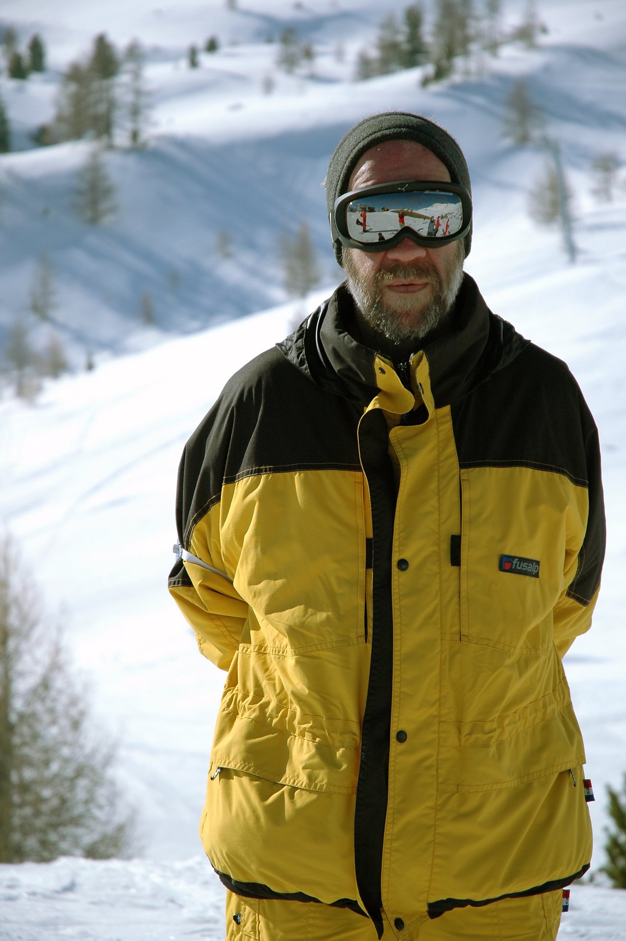 A man in a yellow ski jacket and goggles stands in the snow, with skiers reflected in his lenses.