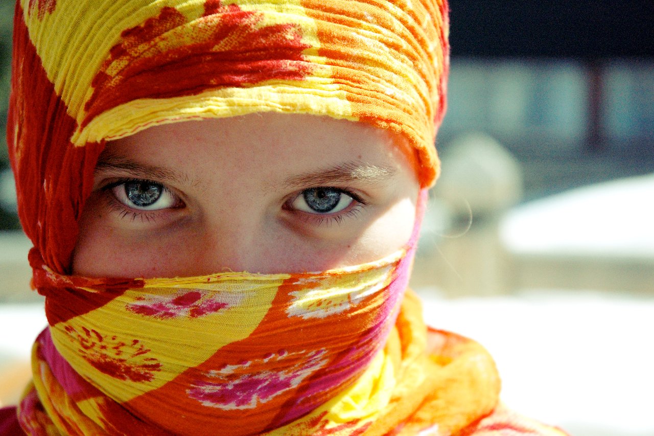 A person with blue eyes wears a colorful headscarf, covering most of their face, and looks directly at the camera.