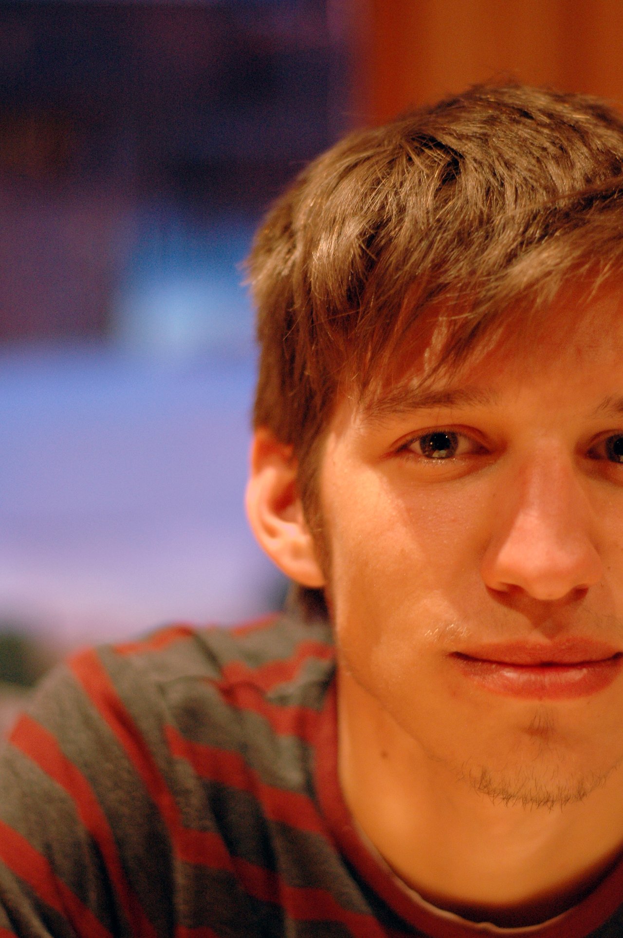 A young man with short brown hair and a striped shirt sits indoors, looking directly at the camera.