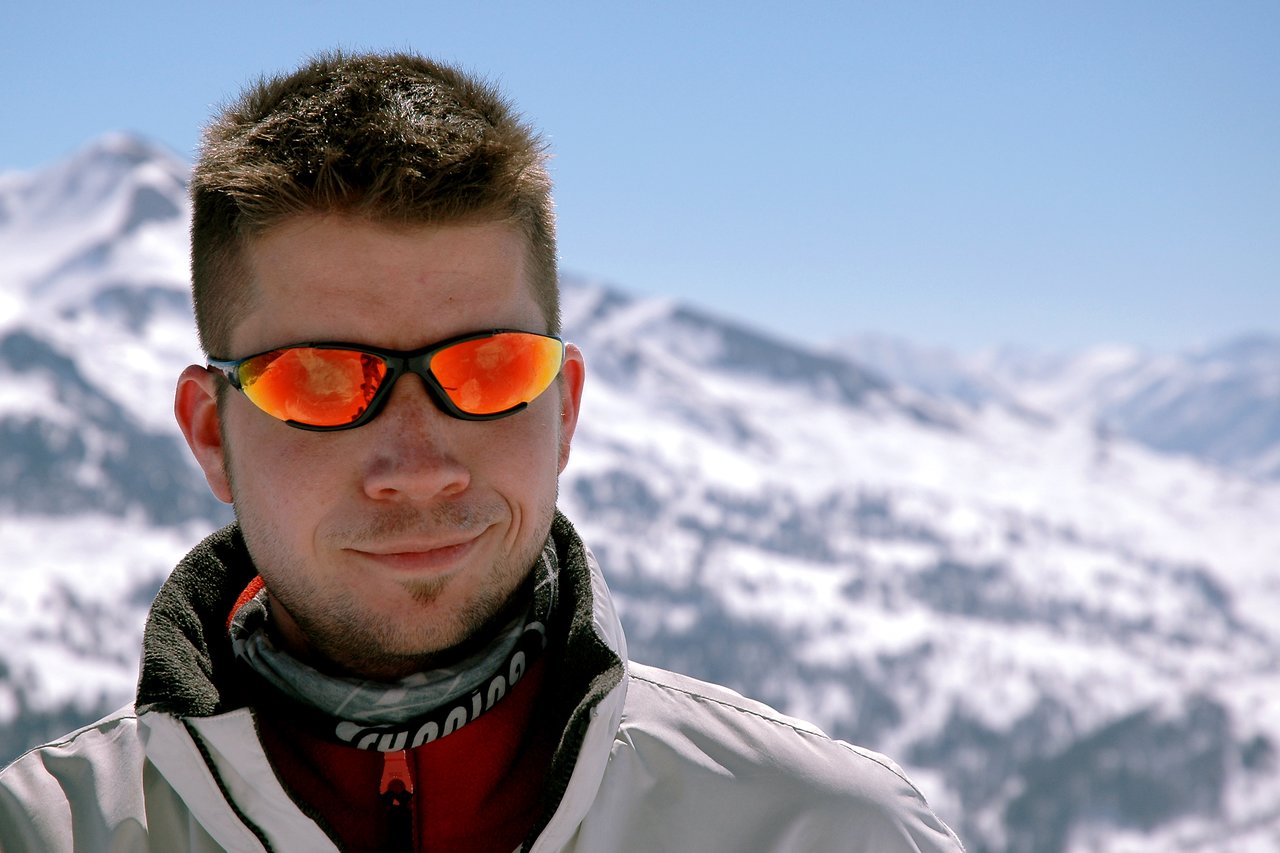 A man wearing reflective sunglasses and a winter jacket stands outside, smiling slightly, with snowy mountains behind him.