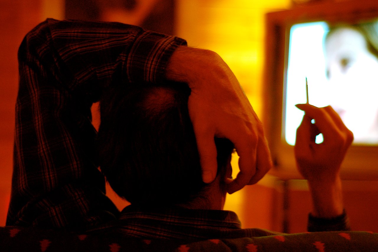 A person sits with one hand on their head and the other holding a toothpick, watching television.