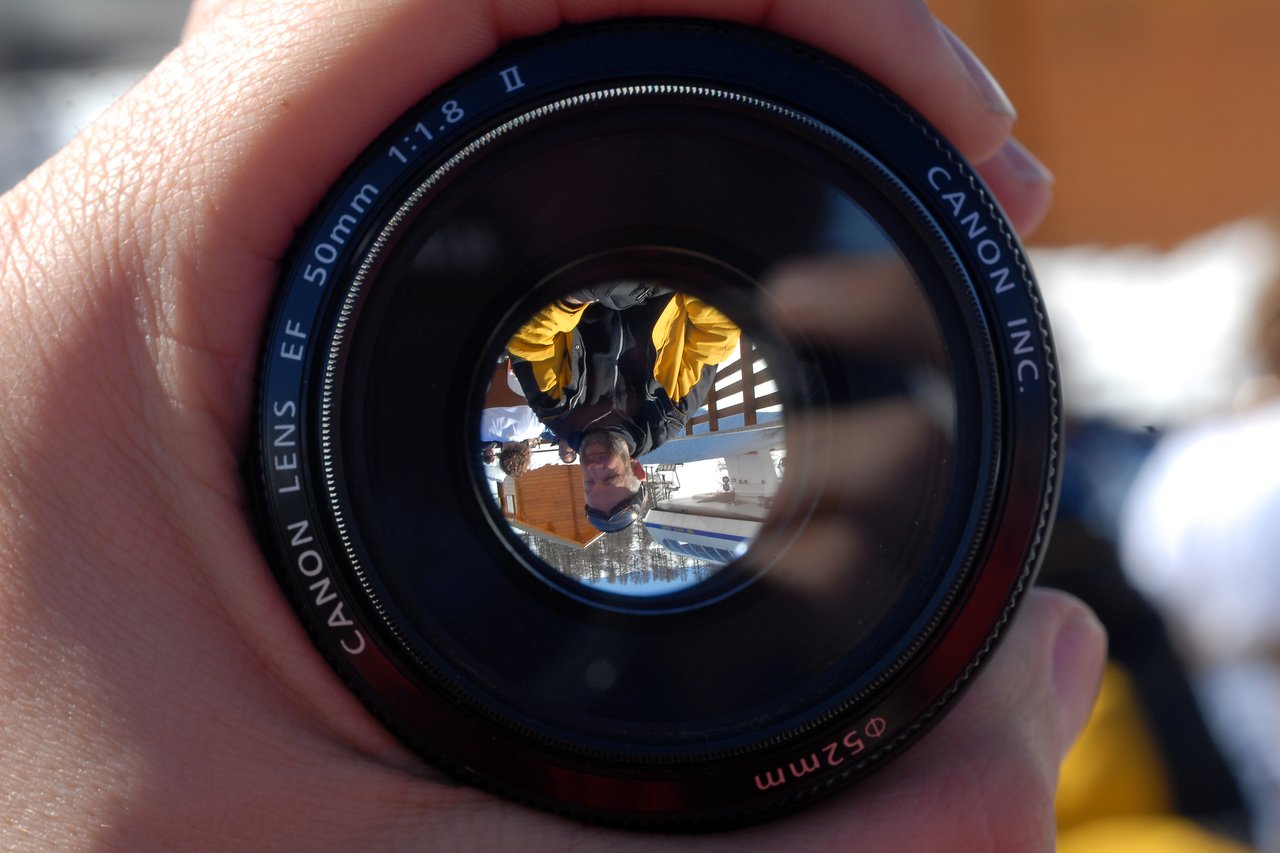 A hand holds a Canon camera lens, showing an upside-down reflection of a person wearing a yellow and black jacket.