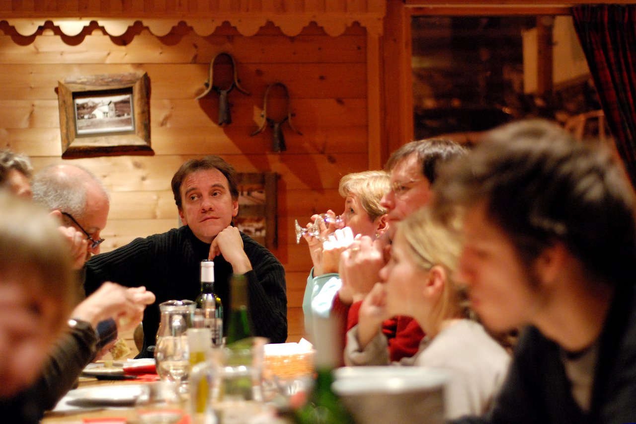 A group of people sits around a table, eating, drinking, and talking in a warmly lit restaurant.