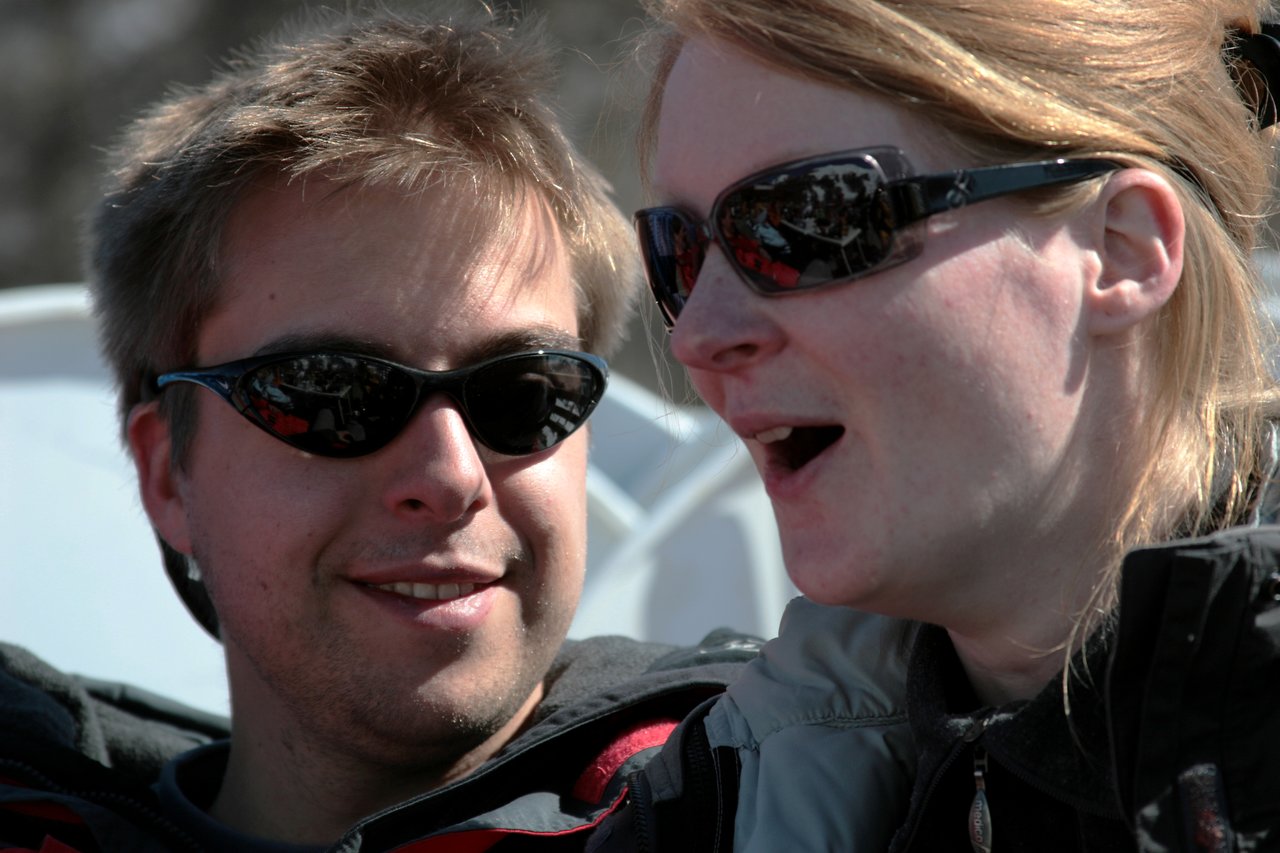 A man and a woman wearing sunglasses sit close together, with the woman speaking or reacting to something.