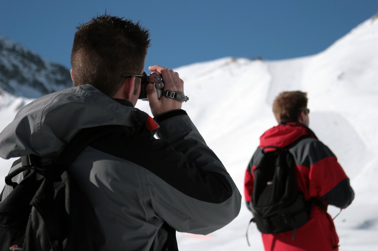 A person in a gray jacket films with a small camera while another person in red looks at the snowy landscape.