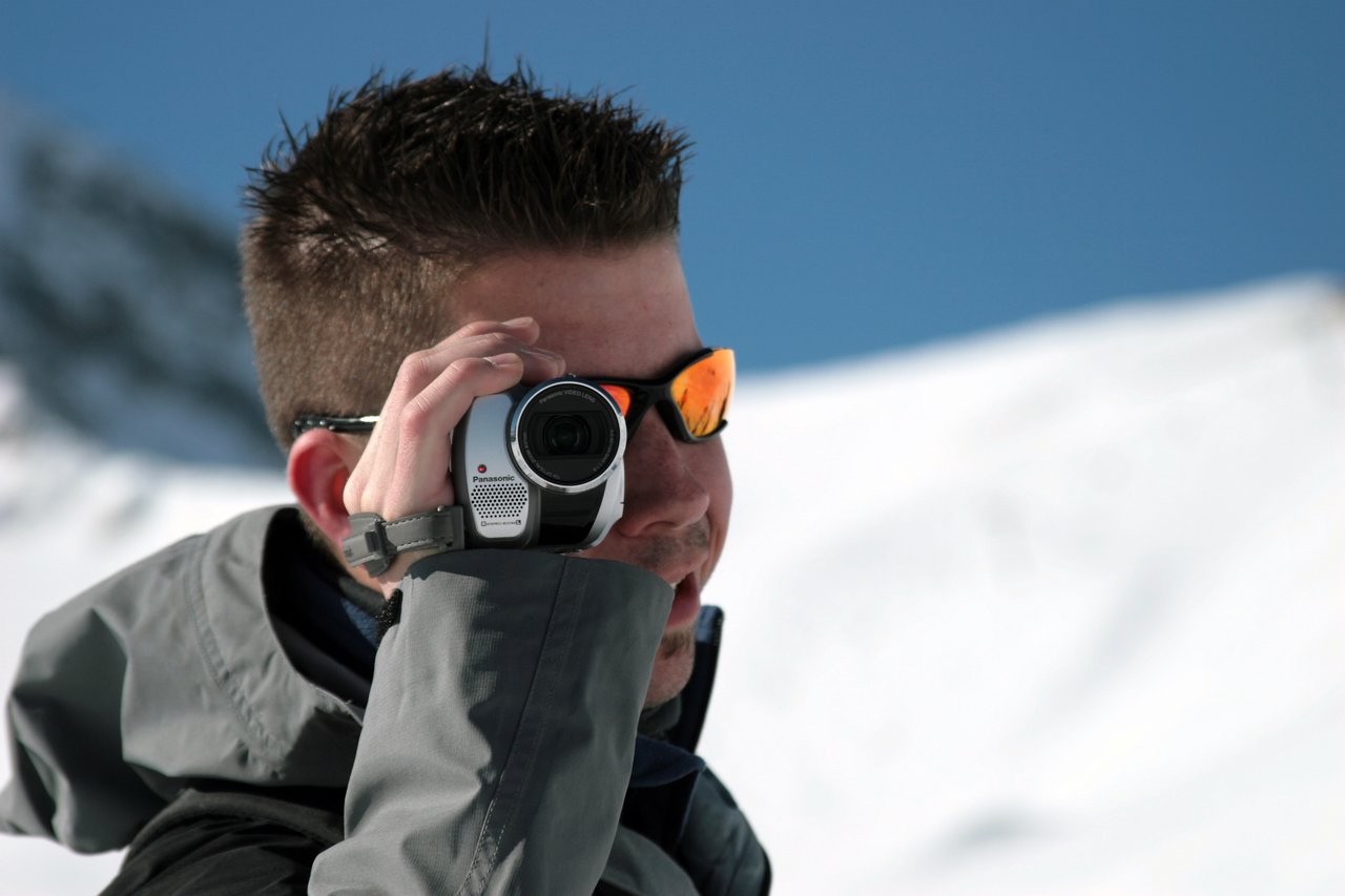A person wearing sunglasses and a winter jacket films with a handheld camcorder in a snowy outdoor setting.