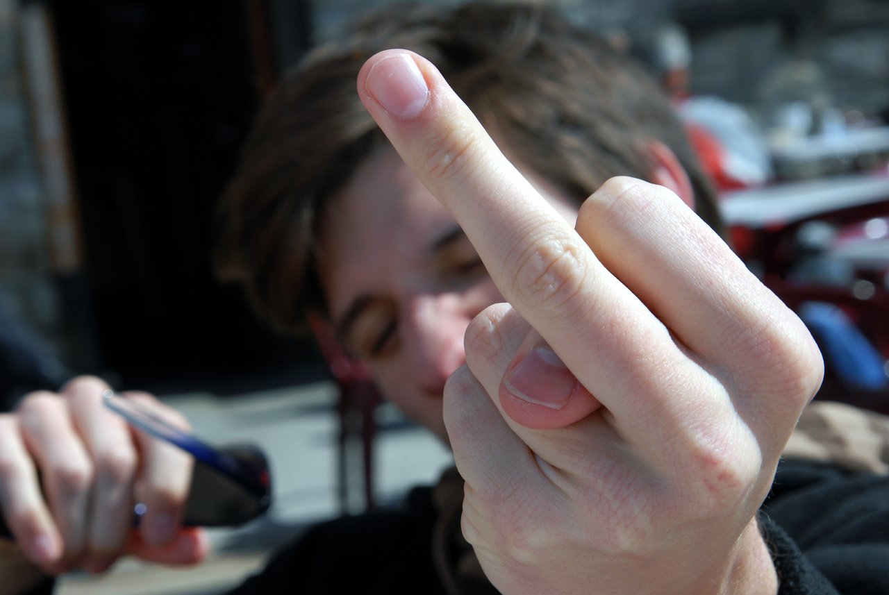 A person holds up their middle finger close to the camera, with their face slightly blurred in the background.
