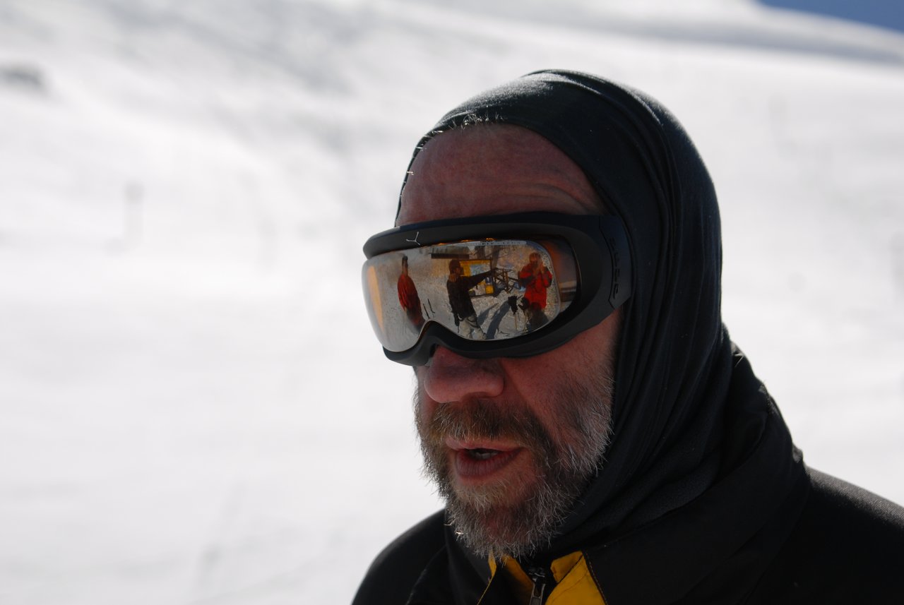 A man wearing ski goggles and a hooded jacket stands in the snow, with people reflected in his lenses.