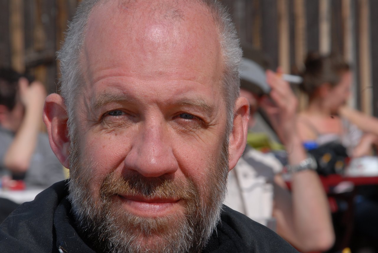 A bearded man with a shaved head looks directly at the camera, with people socializing in the background.