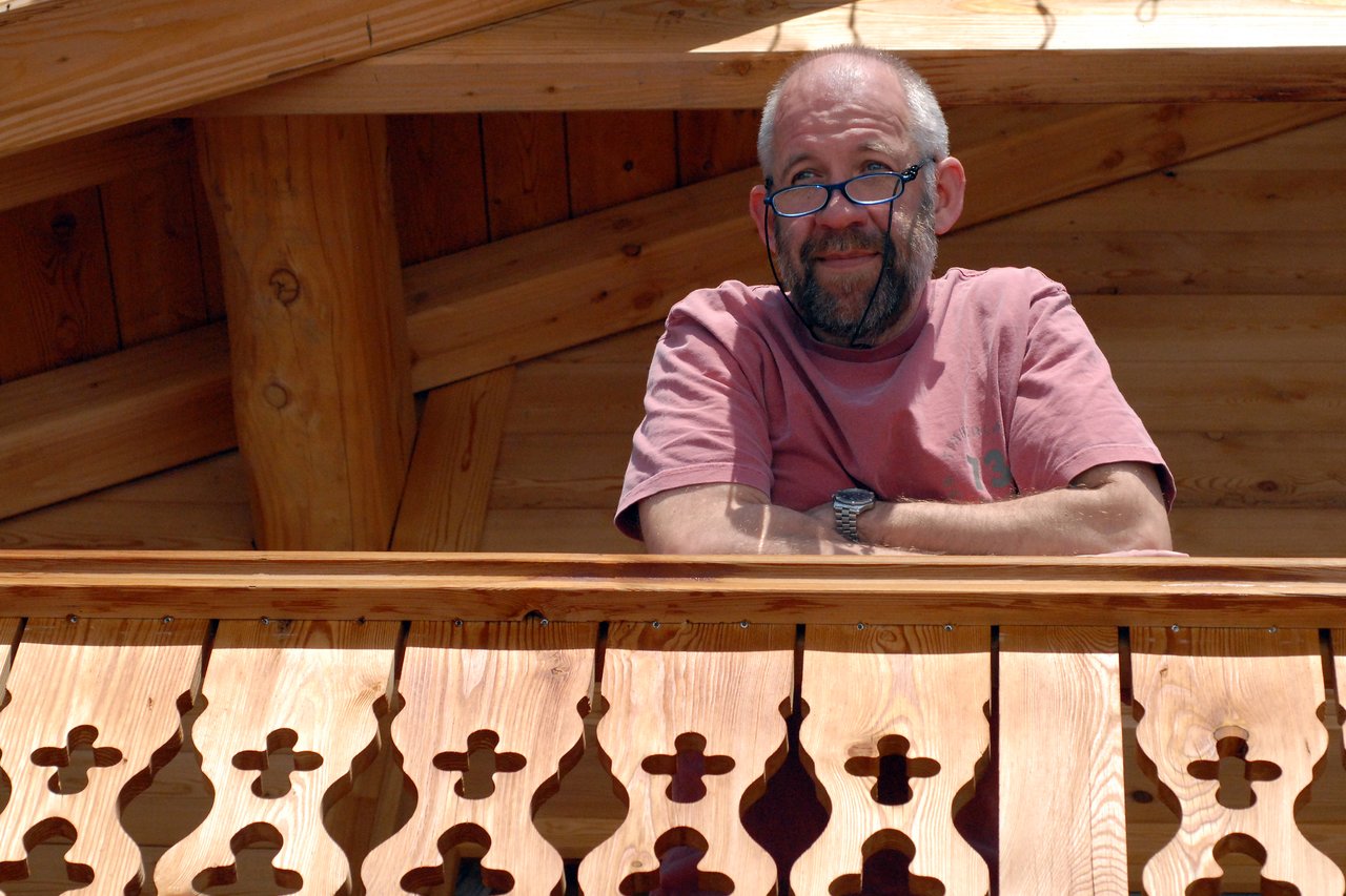 A man with glasses and a beard leans on a wooden balcony railing, looking ahead with a slight smile.