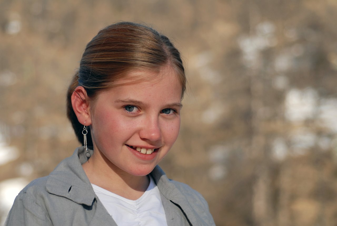 A young girl with light brown hair and blue eyes smiles at the camera, wearing a gray jacket and earrings.