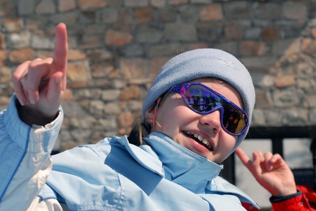 A smiling person wearing sunglasses and a winter jacket points upward with one hand.