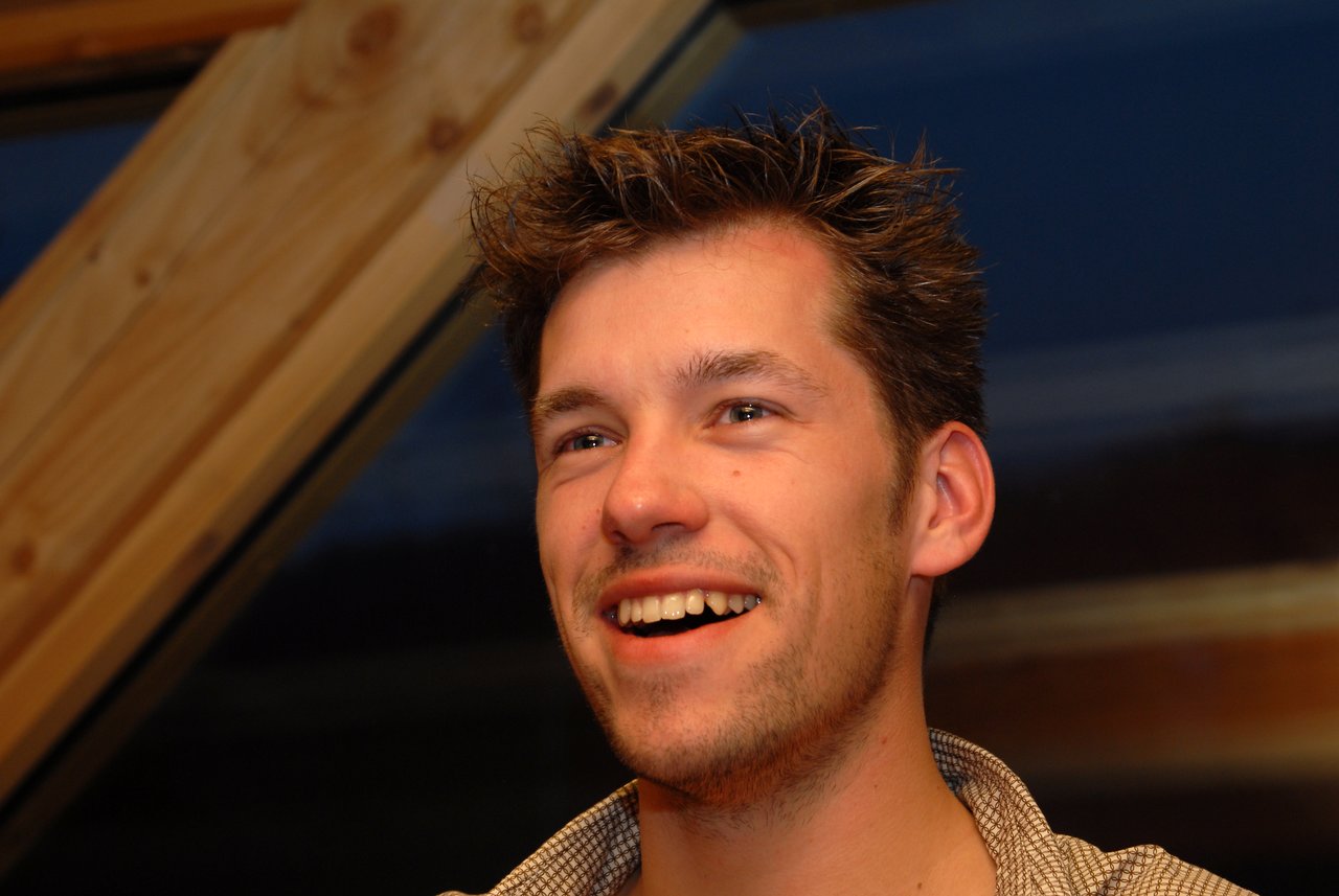 A man with short, spiky hair smiles and looks off to the side in warm indoor lighting.